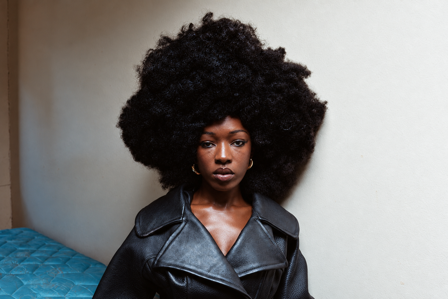 Elsa Majimbo in a black coat, sitting on the edge of a bed, with an afro hairstyle