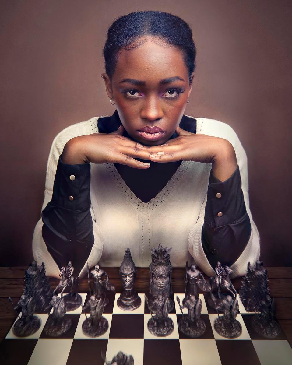 Elsa Majimbo rests her hands under her chin in front of a chessboard with carved pieces