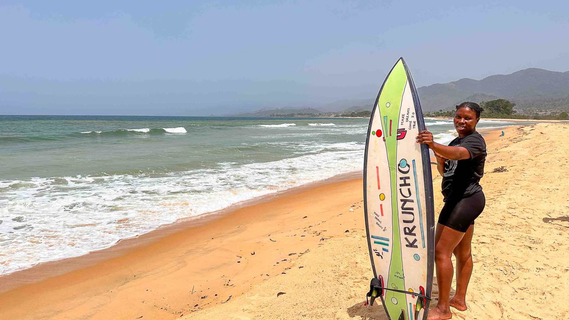 Kadiatu Kamara standing on the beach, holding her surfboard