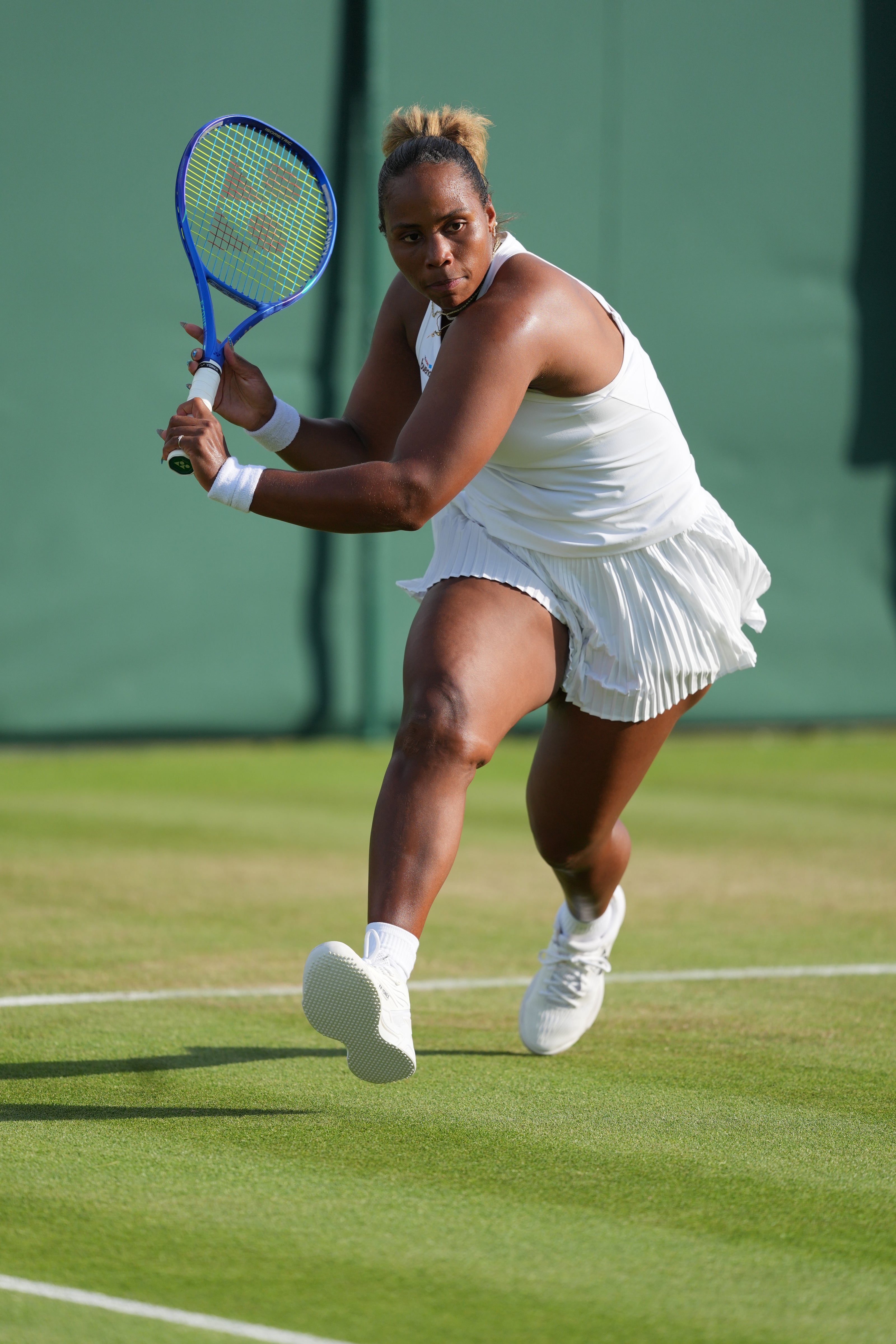 Taylor Townsend, in white, holds a blue racquet in front of a green wall